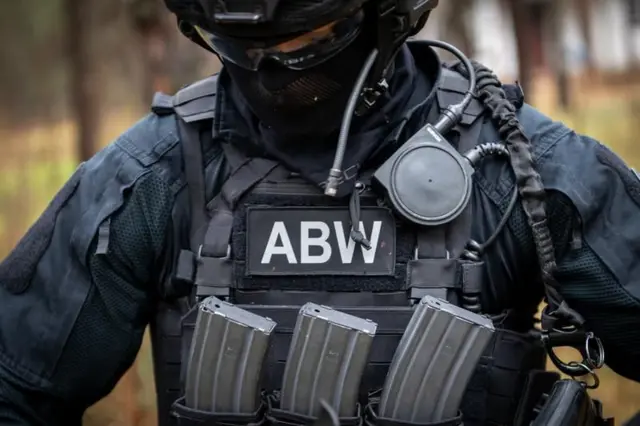 Співробітник польської спецслужби в чорному тактичному спорядженні, шоломі та захисній масці. На бронежилеті розміщена нашивка «ABW» (Агентство внутрішньої безпеки Польщі), а також магазини до зброї та засоби зв'язку.