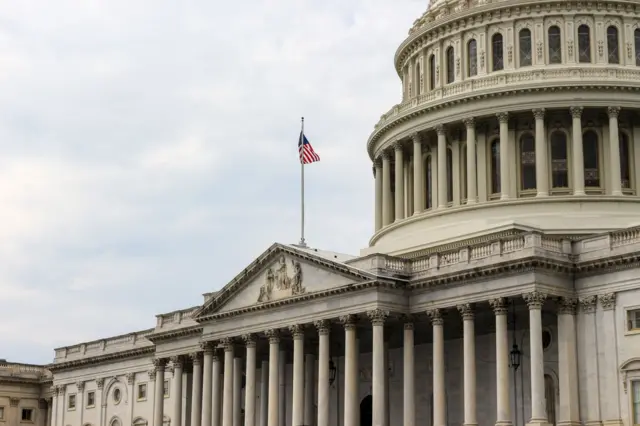 صورة فوتوغرافية من زاوية منخفضة للبيت الأبيض والعلم الأمريكي مرفوعاً عليه، في مواجهة سماء ملبدة بالغيوم.