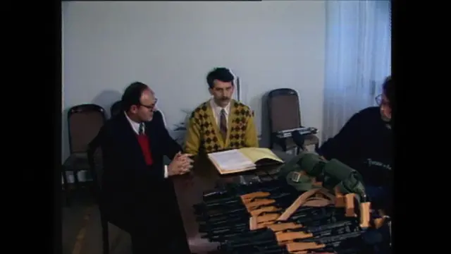 Два мушкарца седе испред велике количине оружја, један има бркове