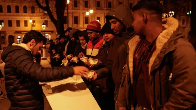 Trieste'deki bir meydanda, palto ve şapkalı bir grup adama paket yemek dağıtan bir adam (Şubat 2024)