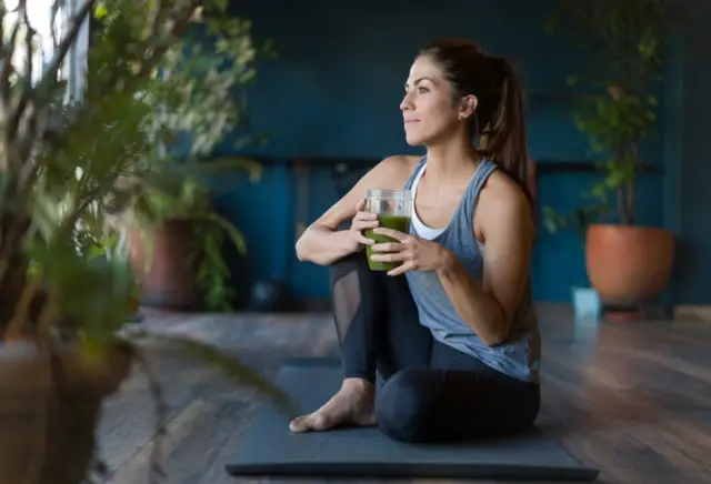 Una mujer joven toma un jugo verde antes de comenzar su clase de yoga 