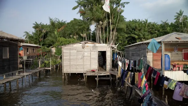 Casasbeta roletamadeira erguidas sobre palafitas, com varais com roupas coloridas à frente, e palmeiras ao fundo