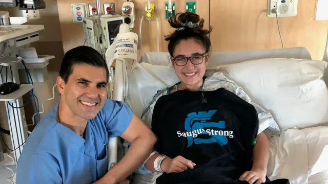 Mia Tretta is in a hospital bed wearing hospital gown with a black top draped over it that says Saugus Strong in white letters. A surgeon sits next to her in blue surgical scrubs. Both of them are smiling. 