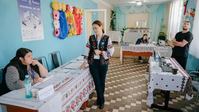Избирательный участок: на стене флаг Молдовы, украшенный цветами, столы покрыты скатертью с традиционным узором, женщина в блузке и жилетке с народными мотивами стоит у стола, за каждым столом сидит по человеку.