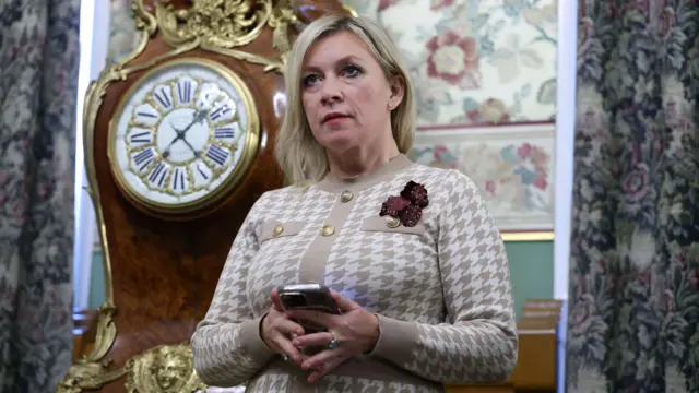 Marija Zaharova stoji u prostoriji isped sata, držeći telefon u rukama