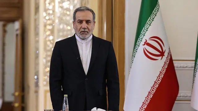 伊朗外交部长阿巴斯‧阿拉格奇立于伊朗国旗旁；他身穿黑色西装外套，内搭一件白色无领波斯式衬衫。