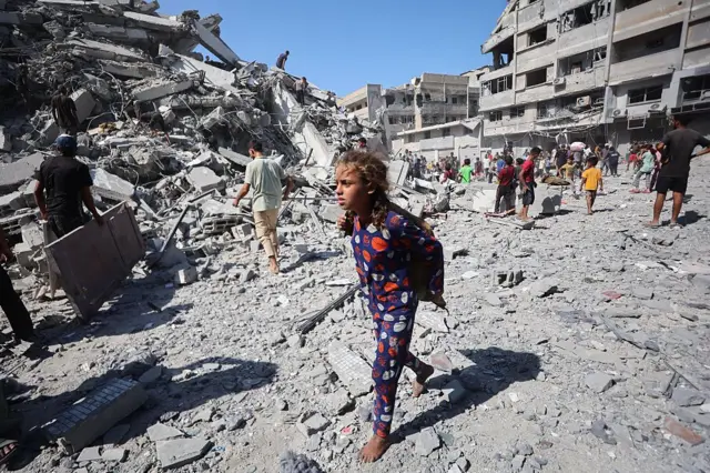 A girl wearing what appear to be colourful blue pyjamas with red and white spots walks across rubble barefoot as displaced Palestinians salvage items from the ruins of the al-Ghafri tower after it was destroyed by Israeli strikes. 