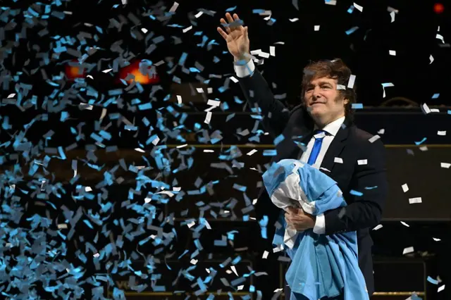Milei saúda o público durante o lançamento de seu novo livro, segurando a bandeira argentina.