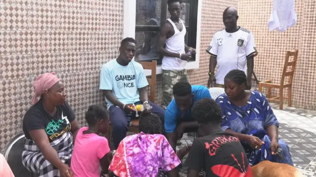 Yankuba Minteh, assis sur une chaise, fait partie d'un groupe de sept personnes vêtues de vêtements aux couleurs vives, assises en cercle pour partager un repas provenant d'un plat commun, qui se trouve hors champ, posé sur le sol. Deux autres hommes se tiennent debout à l'extérieur du cercle et observent la scène. Le repas se déroule à l'ombre, dans une cour carrelée.