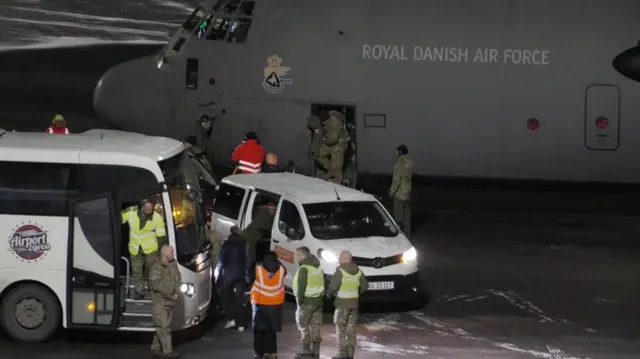 地面近くまで胴体を下げた灰色の機体に「王立デンマーク空軍」と白い文字で書かれた輸送機から、迷彩服姿の兵士たちが降りてきている。手前に、空港内の移動用車両が停まり、蛍光色のベストを着た人たちが、白い氷の見える駐機場に待機している