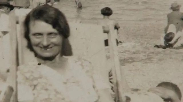 Black and white photo of Louisa Dunne. She is sat on a deck chair looking at the camera and smiling.
