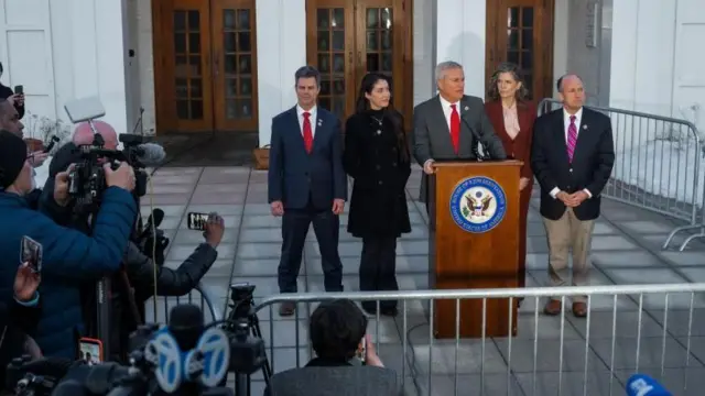 白い壁に、ガラスがはめ込んである茶色い木製のドア3枚を背に、木製の演台に両手をつく背広姿の白髪の男性。左右に男女の議員が計4人並んでいる。手前に銀色の柵があり、その後ろで報道陣がカメラやマイクを構えている