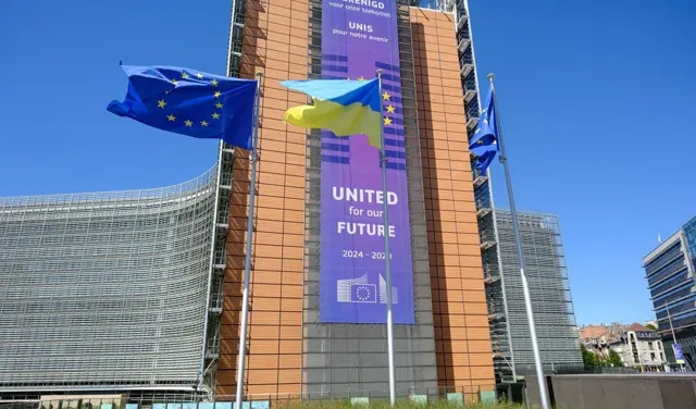 Прапори ЄС та України майоріють на вітрі перед будівлею Берлемон, штаб-квартирою Європейської комісії, з нагоди Дня незалежності України, 24 серпня 2025 року в Брюсселі, Бельгія. На фасаді висить великий банер із написом англійською та французькою: «Об’єднані заради нашого майбутнього, 2024–2029».