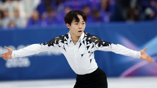 白と黒の衣装の鍵山が両手を広げて滑っている