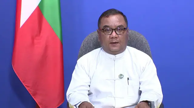 နောက်ခံ အပြာရောင်၊ မြန်မာနိုင်ငံတော်အလံဆက်တင်နဲ့ ရိုက်ကူးရေးအခန်းထဲထိုင်နေတဲ့ တိုက်ပုံအဖြူရောင်ဝတ်ထားတဲ့ စစ်ကော်မရှင်ပြောခွင့်ရ ဗိုလ်ချုပ်ဇော်မင်းထွန်း