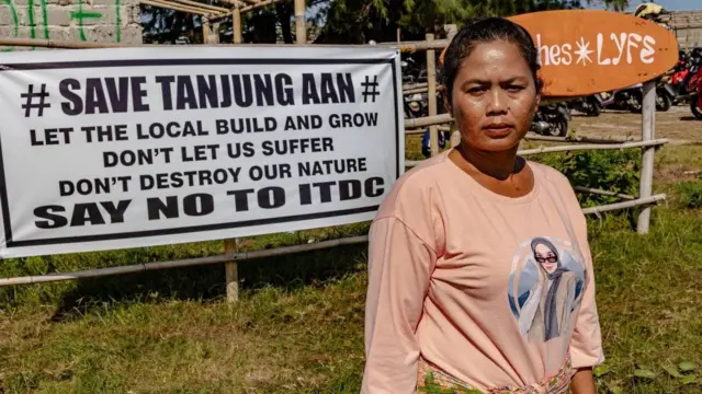 Ella Nurlaila, mengenakan baju lengan panjang berwarna peach, menatap kamera dengan alis berkerut. Di belakangnya, terpampang spanduk besar bertuliskan: "Selamatkan Tanjung Aan, biarkan warga setempat membangun dan menanam, jangan biarkan kami menderita, jangan rusak alam kami, katakan tidak pada ITDC".