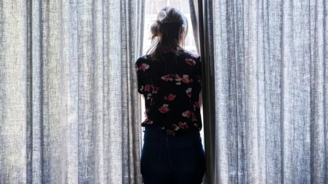 Una mujer, de espaldas a la cámara, mira a través de un par de cortinas grises.
