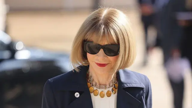 Un plano corto de Anna Wintour luciendo unas gafas negras, un collar dorado y su icónico bob sobre el que recae un rayo de luz