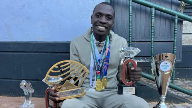 Emmanuel Wanyonyi wears several medals, including his gold from the Paris 2024 Olympics, around his neck as he clutches a Diamond League trophy in his left hand and a World Athletics award in his right hand
