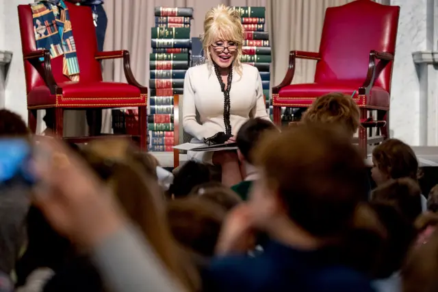 Parton lee su libro "Coat of Many Colors" a los niños, convirtiéndolo en el libro número 100 millones de la Biblioteca de la Imaginación. Lleva gafas de lectura y una camisa beige y marrón. Detrás de ella hay una pila de libros y grandes sillas rojas. 