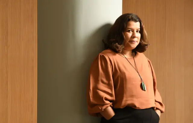 Luciana Chong posa encostada em uma parede marrom com semblante neutro e as mãos nos bolsos da calça preta. Veste uma blusa terracota e usa um colar longo