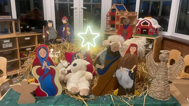 Un belén con corderitos, una estrella iluminada, una mujer rezando, Reyes Magos y dos ángeles. Hay otros juguetes al fondo, sobre muebles que se apoyan contra la pared del fondo y las ventanas.