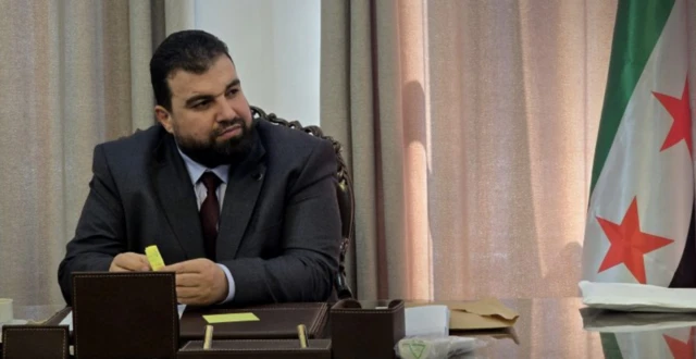 محافظ السويداء، مصطفى البكور يجلس وأمامه مكتب ويرتدي سترة لونها رمادي وقميصاً أبيض وشعره أسود كثيف وله لحية سوداء خفيفة.