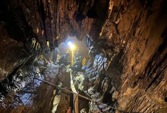 An enormous rocky cavern fills the image with the centre showing the entrance to the cave and four rescuers dressed in blue outfits and helmets using a yellow pump hose, which stretches all the way to the bottom of the picture. A bright yellow light picks out the details of the craggy rocks inside the cave from where they are standing