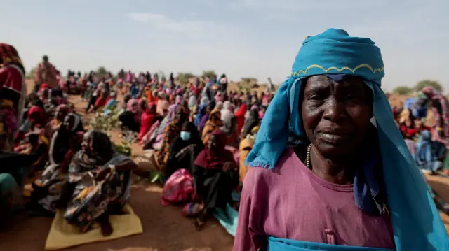 Halime Adam Moussa, mkimbizi wa Sudan ambaye anatafuta hifadhi nchini Chad kwa mara ya pili, akisubiri pamoja na wakimbizi wengine kupokea msaada wa chakula kutoka Shirika la Mpango wa Chakula Duniani (WFP), karibu na mpaka kati ya Sudan na Chad huko Koufroun, Chad, tarehe 9 Mei 2023, huku wakimbizi wengine wakiketi kwenye eneo la wazi nyuma yake.