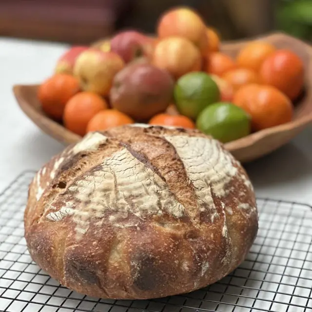 Pão de fermentação natural caseiro sobre uma mesa, ao lado de uma cesta de frutas