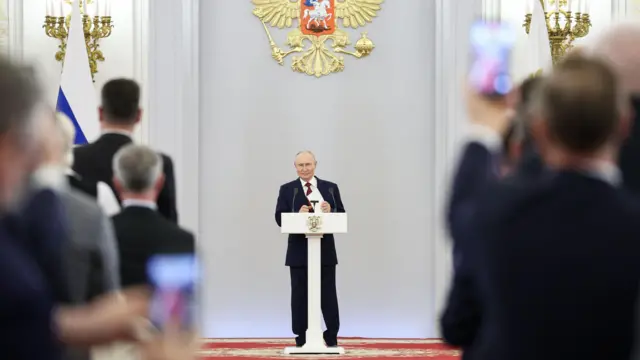 Przemawia prezydent Władimir Putin – mężczyzna w ciemnym garniturze, stojący za białą mównicą.