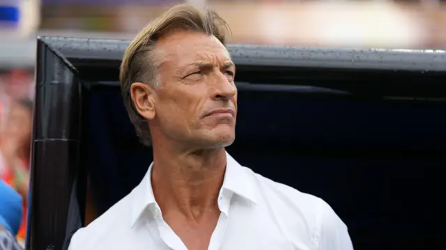 Hervé Renard, dont on aperçoit le buste jusqu'à la tête, vêtu d'une chemise blanche ouverte au col, regarde légèrement sur sa gauche et garde la tête haute, les yeux fixés au loin, debout devant un banc de touche noir dans un stade de football.