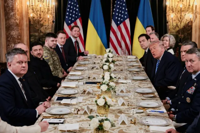 Довгий урочистий стіл, накритий біло-золотистою скатертиною, на якому – порцелянові тарілки з золотим обрамленням, келихи для води та вина, а також композиції з білих троянд у золотих вазах.
За столом одна навпроти іншої сидять українська та американська делегації. У фоні видно чотири великі прапори: два прапори України та два прапори США, розташовані чергуванням. Інтер’єр виглядає розкішно, з мармуровими стінами та декоративними елементами.