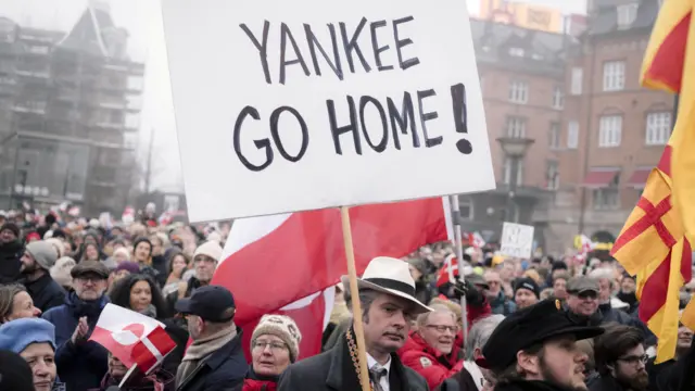 Толпа на на городской площади. На переднем плане - статный усатый мужчина с каменным лицом, в шляпе и при галстуке. Он держит в руках плакат на английском «YANKEE GO HOME!» (Янки, валите домой!). На улице явно прохладно, над толпой развеваются флаги, в том числе, Гренландии
