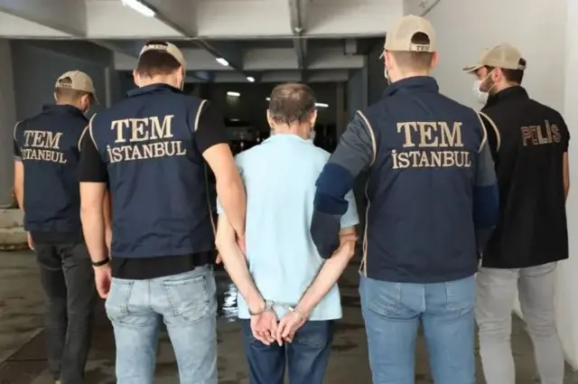 عکسی از چند مرد که پشت آنها به دوربین است، یک نفر مظنون دست بند خورده و مابقی پلیس هستند