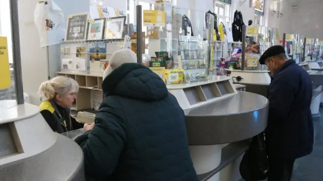 Відділення Укрпошти, чоловік і жінка у зимовому одязі стоять біля віконець обслуговування, спілкуються із працівниками пошти. 