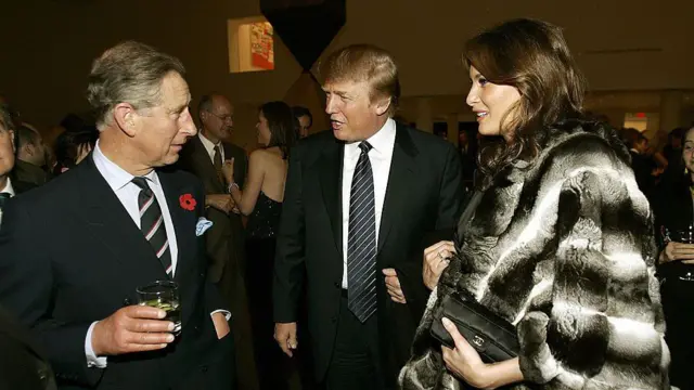 El príncipe Carlos sujeta una bebida mientras charla con Donald Trump, con traje y corbata oscuros, y su esposa, Melania, con abrigo de piel blanco y negro.