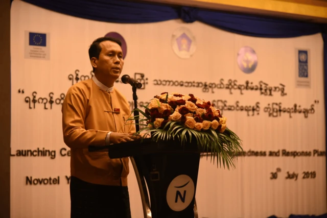 ရန်ကုန်ငလျင်အန္တရာယ် ကြိုတင်ပြင်ဆင်ရေးတုံ့ပြန်‌ရေးစီမံချက်ထုတ်ပြန်ချက်ကို ၂၀၁၉ ခုနှစ်မှာ ပြုလုပ်ချိန် အဲဒီအခမ်းအနားမှာ စင်ပေါ်တက် မိန့်ခွန်းပြောနေတဲ့ ရန်ကုန်တိုင်း ဝန်ကြီးချုပ် ဦးဖြိုးမင်းသိန်း၊ ပင်နီတိုက်ပုံနဲ့ ပုဆိုးကို ဝတ်ဆင်ထားပြီး စင်မြင့်ထက်မှာ စကားပြောနေ 