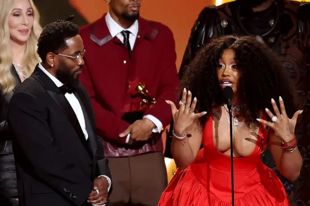 胸元が大きく開いた赤いドレスを着たシザさんが、手を大きく広げながらマイクの前で話している。左隣にはタキシードを着たラマーさんがいる。背後には華やかに着飾った男女が立っている