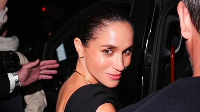 Герцогиня Сассекська повертається до камери, коли збирається сісти в машину. Видно руку, яка супроводжує її. На ній чорна сукня із золотим намистом та сережками-гвоздиками