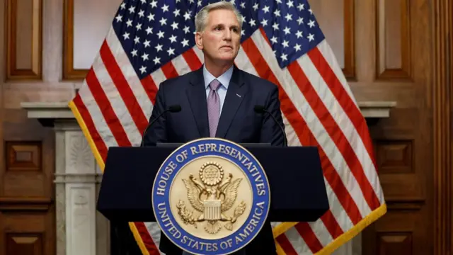 Kevin McCarthy com olhar sério no palanque, que tem símbolo da Câmara dos Representantes e bandeiras dos EUA atrás