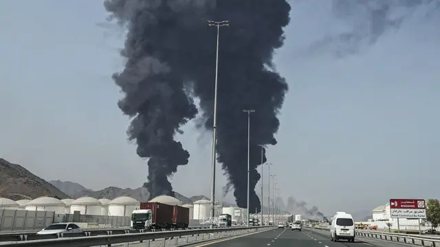 На заднем плане поднимаются в небо два ствола черного дыма. Слева – белые резервуары для нефти. В центре – автомагистраль.