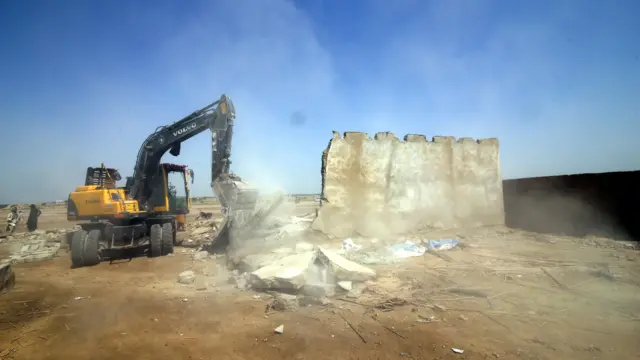 تصویر یک بلدوزر که آوار یک ساختمان ویران شده را جمع می‌کند. یک قسمت از دیوار اردوگاه یا ساختمان هنوز پابرجاست. گرد خاک به هوا بلند شده. آسمان آبی‌است و زمین خاکی