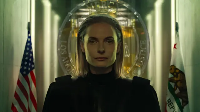 Ребека Фергусон на тамној фотографији гледа у камеру, окружена са две заставе, од којих је једна америчка, иза ње је знак суда