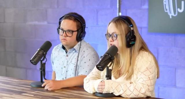 آدری و جرمی، یک زوج جوان، پشت یک میز چوبی نشسته‌اند، هدفون به گوش دارند و مقابل میکروفون صحبت می‌کنند. اتاق حرفه‌ای به نظر می‌رسد و نورپردازی آبی دارد