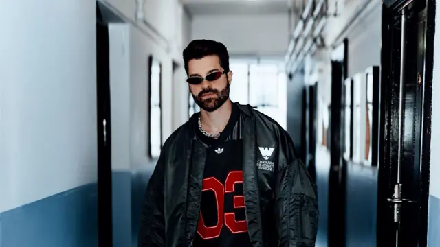 Alok, um homem de barba e cabelo escuro, posa em um corredor de paredes brancas e azul-acinzentadas, olhando diretamente para a câmera com expressão séria. Ele veste um casaco oversized preto sobre uma camisa esportiva preta com o número "03" em vermelho, além de óculos de sol estreitos e um colar prateado, compondo um visual de inspiração urbana e contemporânea.