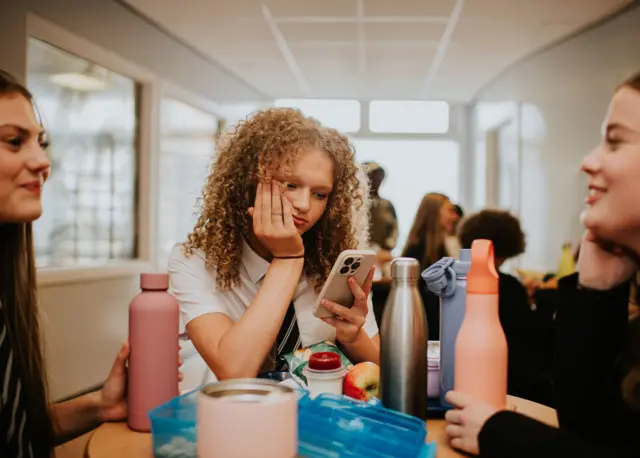 Trzy uczennice siedzą przy stole na szkolnej stołówce. Dwie uśmiechają się i wydają się pogrążone w rozmowie. Trzecia dziewczyna, siedząca pośrodku, patrzy w ekran telefonu. 