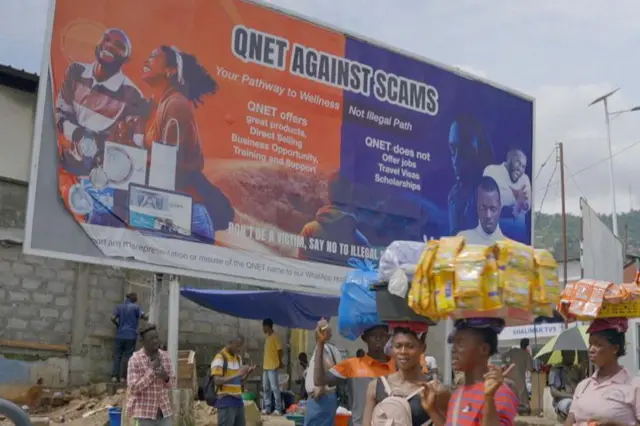 Comerciantes con mercancías en equilibrio sobre sus cabezas pasan frente a una gran valla publicitaria en Freetown, Sierra Leona, en la que se lee "QNET contra las estafas".