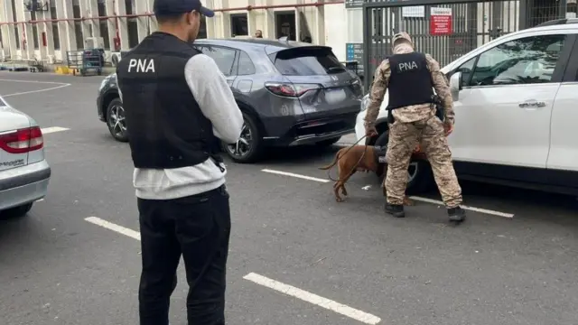 迷彩服姿の隊員がリードをもち、つながれた犬が車のタイヤをかいでいる。少し離れた場所で別の男性がそれを見守っている。