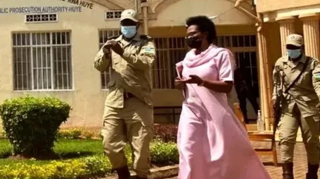 Umugore uri mu mwambaro w'iroza arimo aratambuka akikijwe n'abapolisi babiri